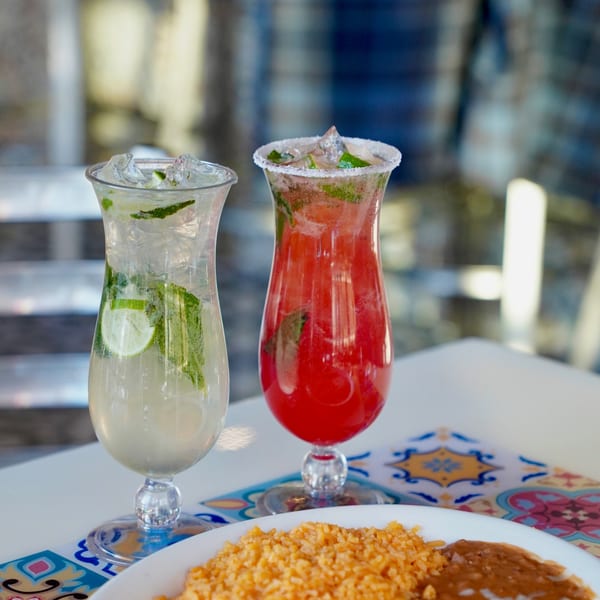 Two vibrant cocktails, one green and one red, with ice and mint, next to a plate of rice and beans.