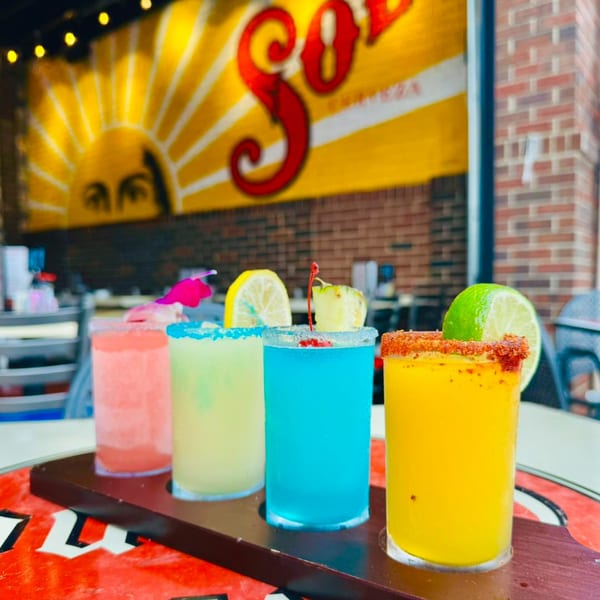 Four colorful margarita shots on a wooden tray, with a "Sol" sun logo in the background.