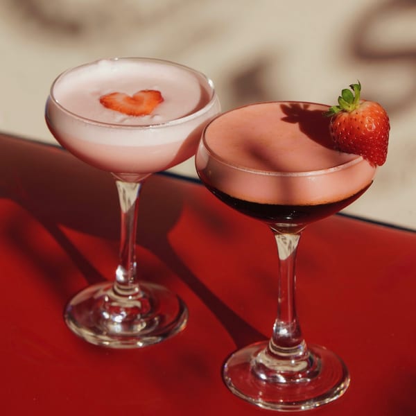 Two strawberry cocktails, one with a heart-shaped strawberry, on a red surface with shadows.