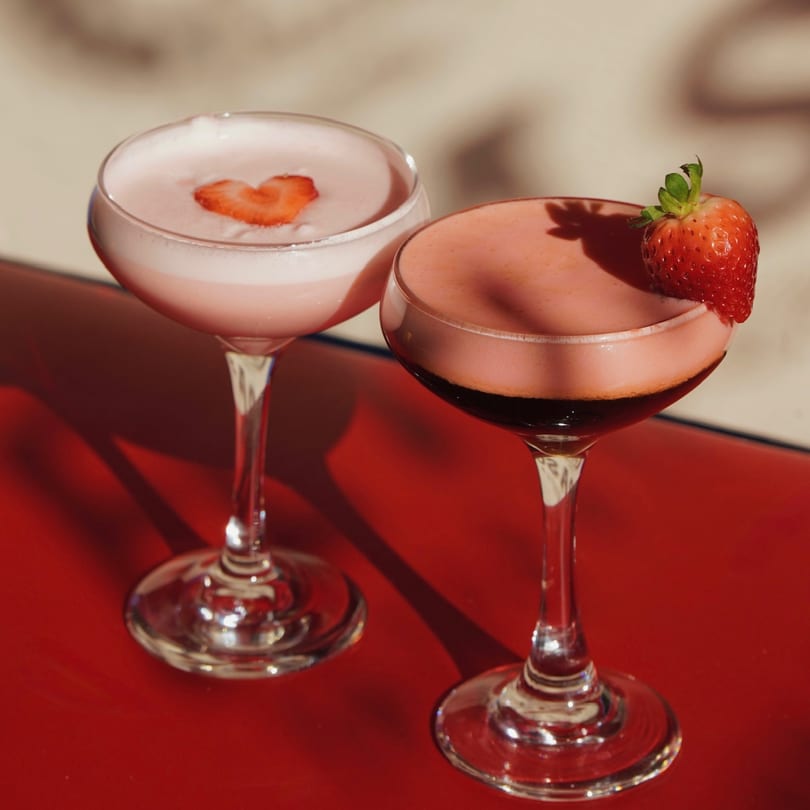 Two strawberry cocktails, one with a heart-shaped strawberry, on a red surface with shadows.