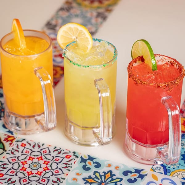 Three colorful margaritas in glass mugs with fruit garnishes and patterned coasters.