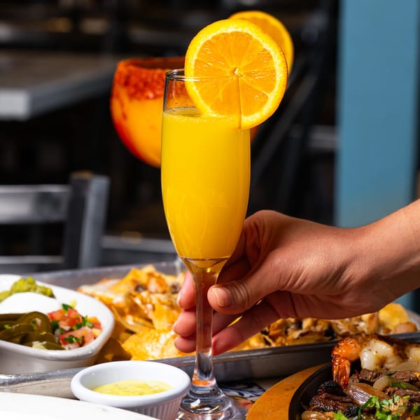 Hand holding a mimosa with an orange slice, surrounded by nachos, salsa, and fajitas.