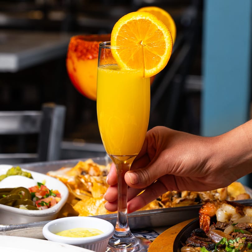 Hand holding a mimosa with an orange slice, surrounded by nachos, salsa, and fajitas.