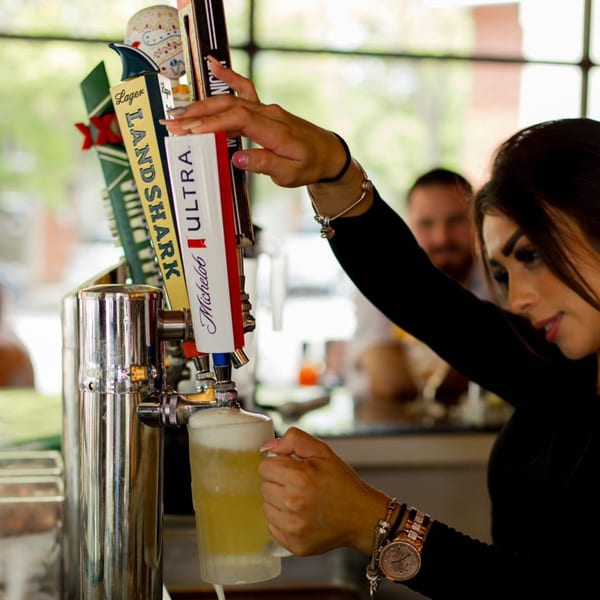 Bartender pouring a frothy beer from a tap into a mug at a lively bar.