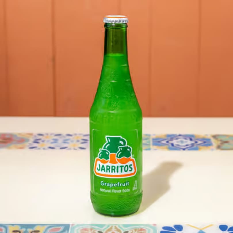 A green Jarritos grapefruit soda bottle stands on a white table with colorful tiled trim.