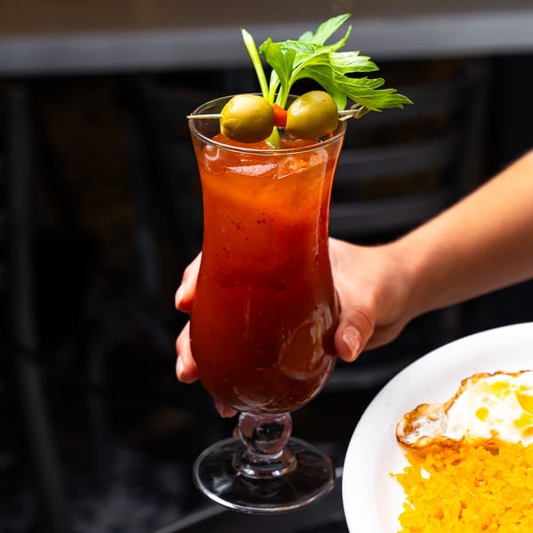 Hand holding a Bloody Mary cocktail garnished with olives and celery, next to a plate of rice and fried egg.