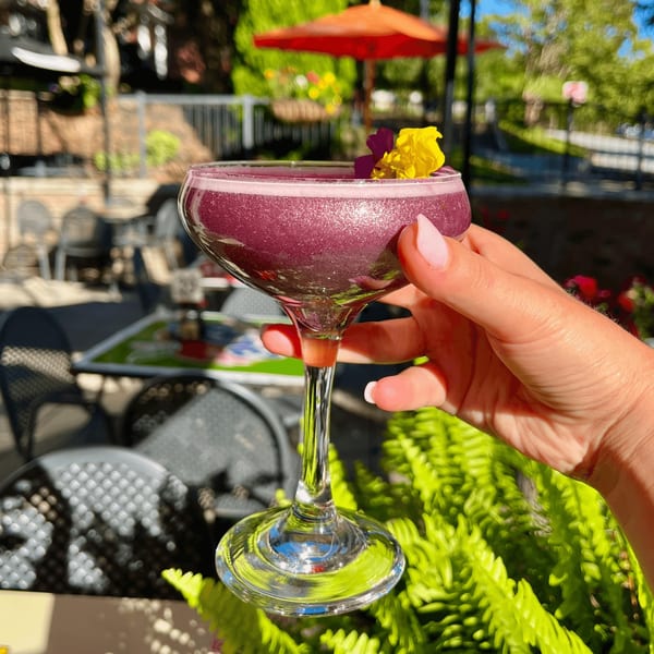 A hand holds a sparkling purple cocktail garnished with yellow and purple flowers on a sunny patio.