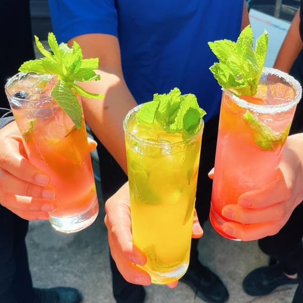 Three hands hold colorful cocktails garnished with fresh mint, one with a sugared rim.
