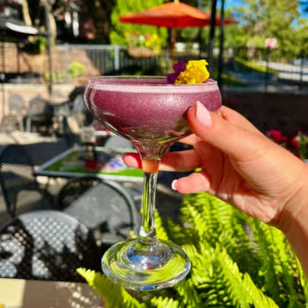 Hand holding a purple cocktail garnished with yellow and purple flowers on an outdoor patio.