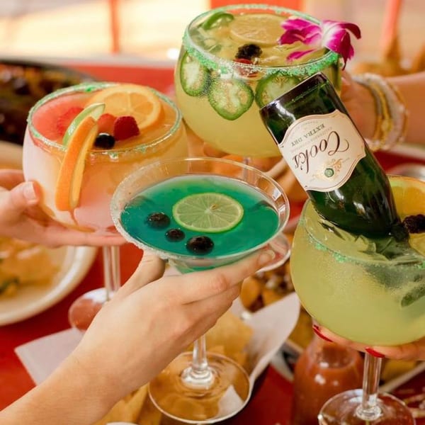Four hands holding colorful margaritas, one with a mini champagne bottle, on a red table.