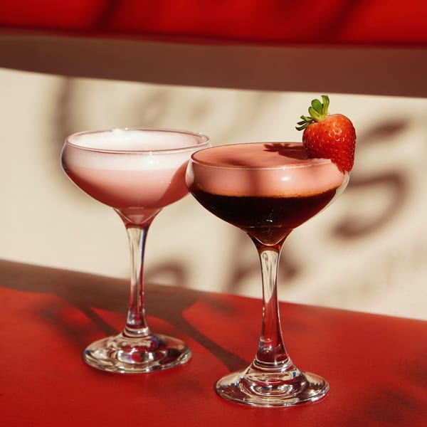 Two pink cocktails, one garnished with a strawberry, sit on a red surface in sunlight.