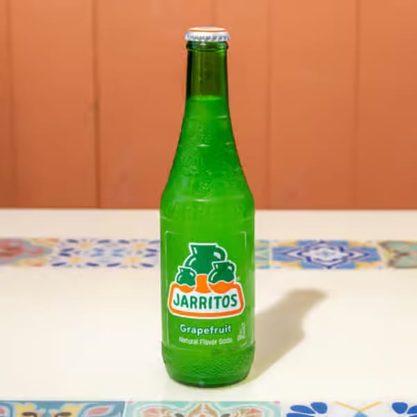 A green glass bottle of Jarritos Grapefruit soda stands on a white table with colorful tiles.