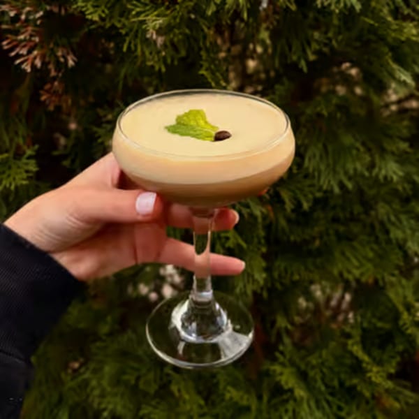 Hand holding a creamy cocktail garnished with a mint leaf and coffee bean against green foliage.