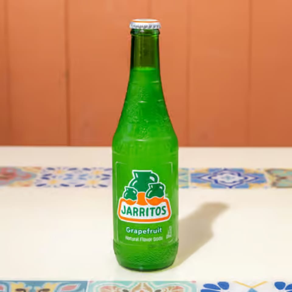 A green Jarritos grapefruit soda bottle stands on a white table with colorful tiled trim.