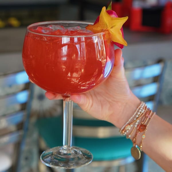 A hand holding a large glass of red cocktail garnished with a star fruit slice and pink flower.
