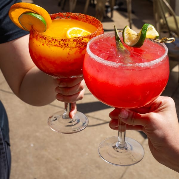 Two hands holding vibrant red and orange margaritas with fruit garnishes, clinking glasses outdoors.