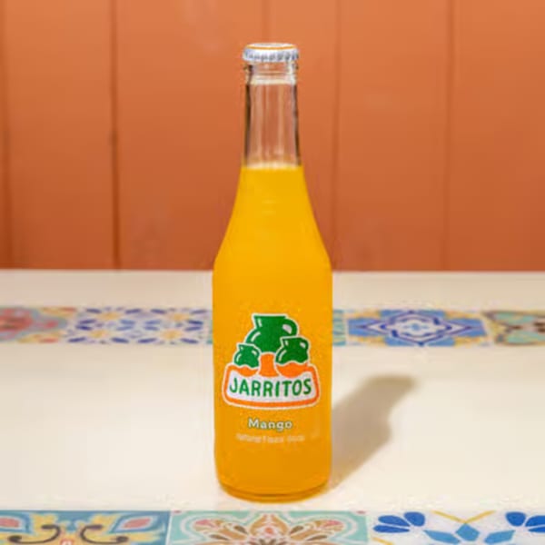 A bottle of Jarritos Mango soda with a silver cap sits on a colorful tiled surface.