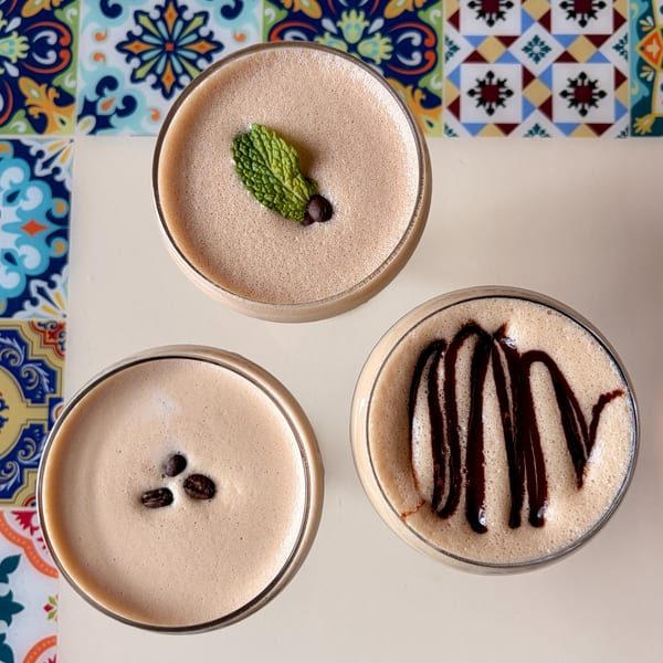 Three espresso martinis with foam, one garnished with mint, one with coffee beans, and one with chocolate drizzle.