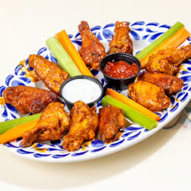 A platter of spicy chicken wings with celery, carrots, ranch, and hot sauce on a white and blue plate.