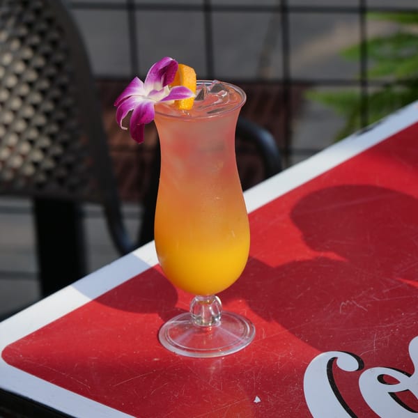 A vibrant orange and pink cocktail in a hurricane glass, garnished with an orchid and orange slice.