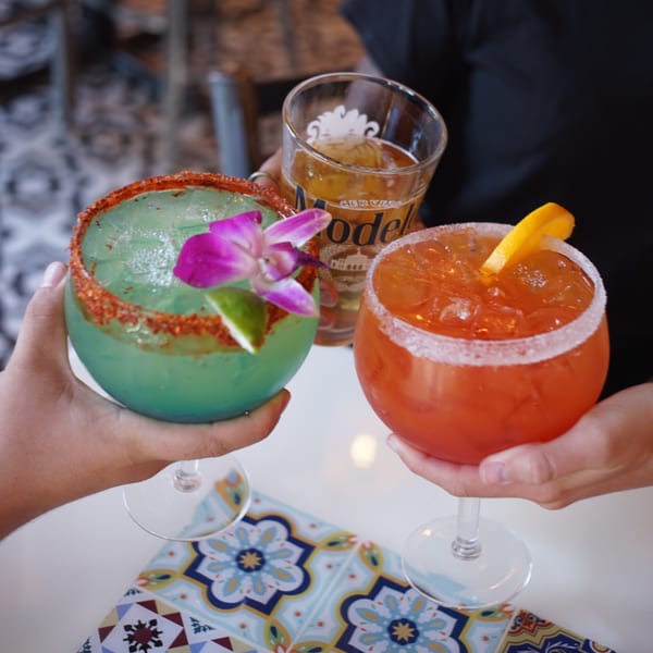 Three hands clinking a green margarita, an orange margarita, and a beer.