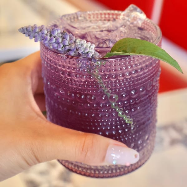 Hand holding a purple cocktail in a textured glass, garnished with lavender and a green leaf.